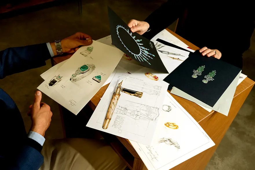 Two people working at a desk with sketches of jewellery and green gemstone